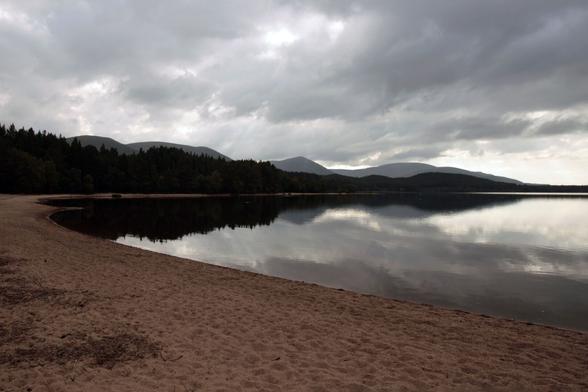Dunkle graue Wolken spiegeln sich in einem See. Im Vordergrund braun-roter Sandstrand, im Hintergrund Berge und Wälder. Die Farben sind eher gedeckt, die Stimmung ruhig.

Dark gray clouds are reflected in a lake. Brown-red sandy beach in the foreground, mountains and forests in the background. The colors are rather muted and the mood is calm.