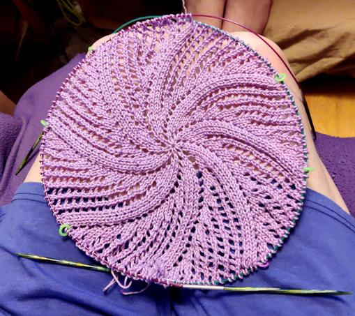 A knitted doily worked in the round on 2 circular needles. The spiraling pattern has become evident. It remains a shade of lilac