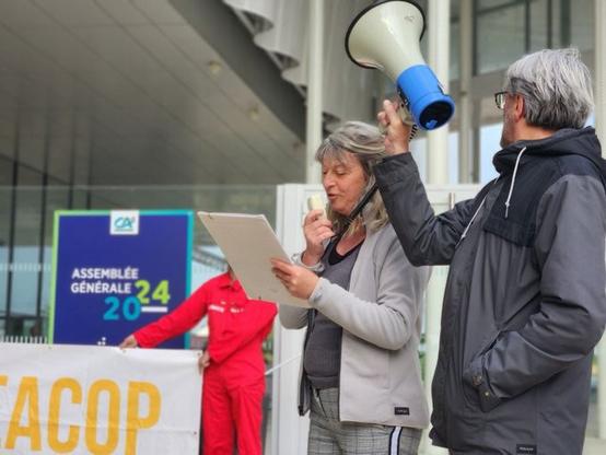 Une militante prend la parole dans le micro d'un mégaphone, tenu par un autre militant. Les deux sont de profil. 
Derrière eux, un militant tient le bout de la banderole Stop EACOP. Tout au fond, l'affiche de l'assemblée générale.