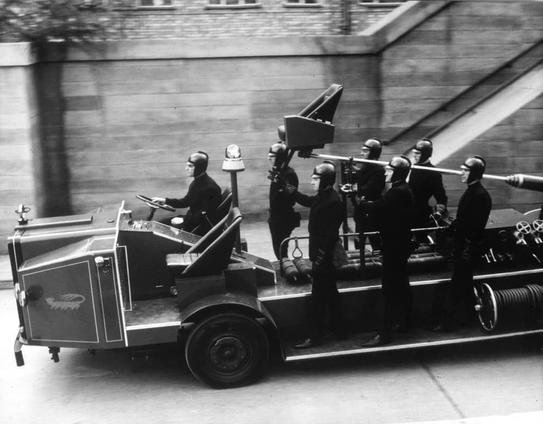 The image depicts a scene from the movie "Fahrenheit 451." In the foreground, there is an old-fashioned fire truck with two people riding on top. They appear to be part of a crew involved in some sort of emergency or action sequence. On the back of the truck, there are several other individuals dressed in similar uniforms, suggesting they might also be part of the same crew or organization. The truck is driving down what seems to be a busy street with various buildings and infrastructure visible in the background. There are no clear texts within the image.
