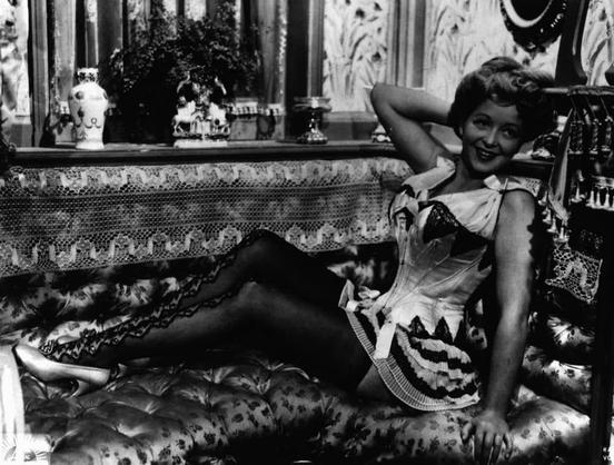The image depicts a woman sitting on a couch. She is wearing a dress with a high slit, revealing her legs. Her pose suggests a sense of relaxation or casualness. There is no visible text in the image. The style and quality of the image indicate that it could be from an older film, given its grainy texture and black-and-white tone.