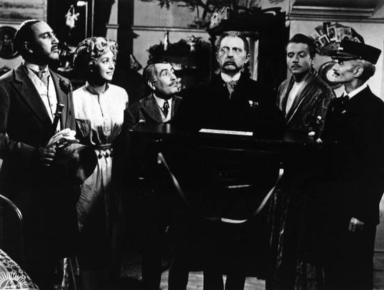 The image appears to be a still from a film, showcasing a group of people gathered in what looks like an old-fashioned setting. There are at least eight individuals visible, with various attire suggesting different characters or roles within the scene. One person is seated and others are standing, creating a sense of conversation or interaction among them.

The lighting and composition suggest it's a black and white film, which is typical for films from earlier times or those stylized to look like they were made in a specific era. The clothing and hairstyles also hint at the period setting, possibly Victorian or early 20th century based on the style of costume.

The title "Autore non identificato - Danielle Darrieux in 'Occupati d'Amelia'" suggests that this image may be from a film featuring Danielle Darrieux and is associated with an unidentified author or director, adding to the sense of intrigue about the context of this scene.