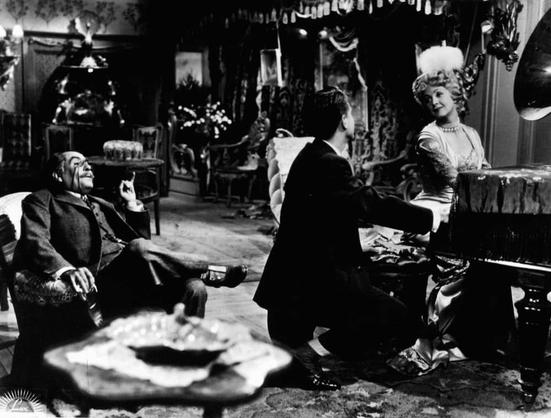 The image appears to be a still from an old black and white movie. It features two people, a man and a woman, in what seems to be a living room setting with antique furniture. The man is seated comfortably on a sofa, smoking a pipe and looking at the woman with a relaxed posture. The woman stands next to him, facing away from the camera and towards the man, seemingly engaged in conversation or waiting for her turn to speak.

There are indications of a festive atmosphere, such as a decorated Christmas tree in the background, which adds a touch of warmth and holiday cheer to the scene. The overall mood of the image is nostalgic and intimate, capturing a moment from a bygone era of cinema.

Please note that this description is based on the available visual information and does not include any speculative or inferred details.