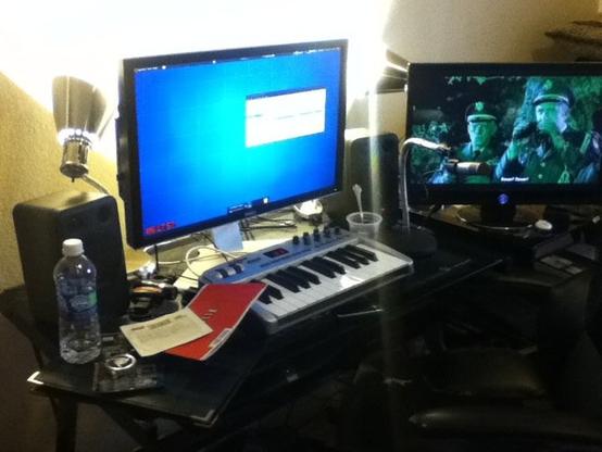 A glass desk with a dual-monitor computer setup, a couple of large PC speakers by M-Audio, an empty water bottle, a small MIDI keyboard, and a Netflix envelope. On the L monitor we see an Ubuntu desktop background, while a movie plays on the R monitor.