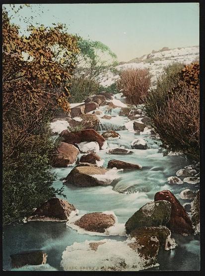 The image depicts a serene natural landscape featuring Gungarline Creek, which is part of the Snowy River in New South Wales, Australia. The stream appears to be flowing gently between steep rock formations and is framed by lush greenery on either side. There are hints of snow visible on the higher ground, suggesting a cool or high-altitude environment. The overall mood of the image is peaceful and picturesque, capturing the natural beauty of this remote area.