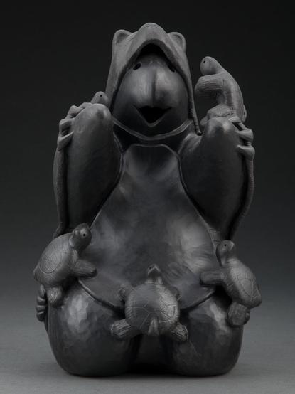 official auction photo of the object, front view on gradient grey-black background: blackware ceramic sculpture depicting a turtle storyteller (sitting upright with mouth open as if speaking) dressed in a bear hide, surrounded by small turtle children listening to it talk