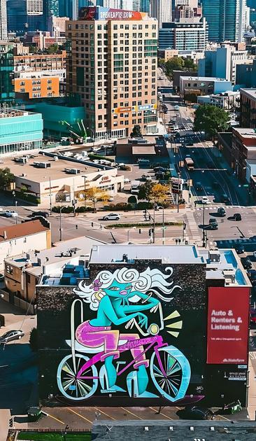 Streetartwall. The mural of a cyclist was sprayed/painted in pop art style on the exterior wall of a multi-storey residential building. The background is black. The girl is painted in turquoise, white, yellow and pink. She is sitting on her turquoise bicycle and has long, white, flowing hair with flowers. The bike is reminiscent of a Bonanza bike (or "High Riser Bike"), which looks a bit like a chopper that is too short. She is wearing pink trousers, has a turquoise bare upper body and is wearing white high heels. She is casually leaning on the handlebars, and since the bike light is on, she seems to want to ride through the night. (The photo shows a large red advertising sign on the house and a sweeping view of the surrounding city).