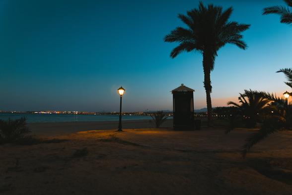 The image depicts a beachfront scene at dusk. A palm tree stands prominently on the sandy beach, with a light fixture nearby and a fence running along the edge of the beach. In the
background, there is a clear view of the ocean with boats visible in the distance. The sky features a beautiful gradient of a late sunset with colors from dark orange to a deep turquoise.