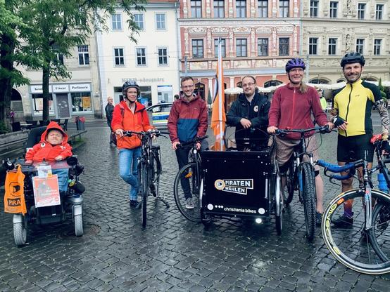 Das Bild zeigt die 6 im Text beschriebenen Kandidaten, Markus Walloschek im Rollstuhl, Erik Limburg mit Lastenrad, die anderen mit normalen Fahrrädern. Im Hintergrund die historischen Bürgerhäuser am Erfurter Fischmarkt.