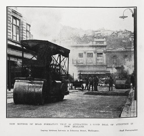 Black-and-white photo: New method of road formation that is attracting a good deal of attention in New Zealand. 1924. Extended caption: Laying bitumen hot-mix in Johnston Street, Wellington. Description: Shows a steamroller at left foreground and another at right background; and workmen spreading bitumen on the road surface. Two-storey buildings surround the street, seen at left and behind the road works. In the far background is a hillside with buildings. Citation: Auckland Weekly News, 2 October 1924, p. 48. Auckland Libraries Heritage Collections AWNS-19241002-48-05. https://kura.aucklandlibraries.govt.nz/digital/collection/photos/id/245817