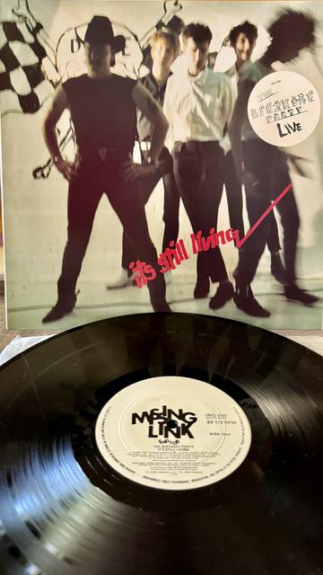 Front cover. Picture of the band, blurred by a motion effect. A white circular sticker says "The Birthday Party Live"

Red text on off white background, overlaid on band picture, reads "its still living"

Below is 12" black vinyl disc with white label with large black letters that read "Missing Link"