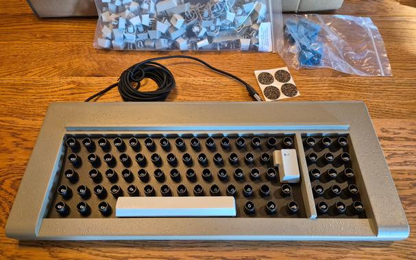 A grey F77 buckling spring IBM repro keyboard on a wooden table, with only the space bar and the iso return key mounted. The rest of the keycaps are in plastic bags behind it, and four cork feet for mounting underneath are also visible.
