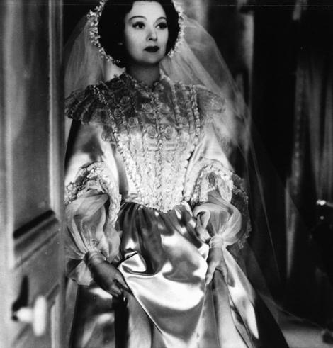 The image depicts a person standing in a hallway. This individual appears to be wearing period attire, consisting of a long dress that extends past the hem of the photo and what seems to be a veil or headdress, suggesting a formal event or historical context. The person is positioned centrally within the frame, with their body facing towards the camera but their head turned slightly away from it, creating an air of mystery or reflection.

The attire and setting suggest that this might be a scene from a film or play set in an earlier time period. However, without further context, such as text or other visual cues, it's difficult to provide more specific details about the person's role, the storyline, or the exact nature of the event they are attending. The style of the image and the quality suggest it might be from a classic film or a historical drama production.