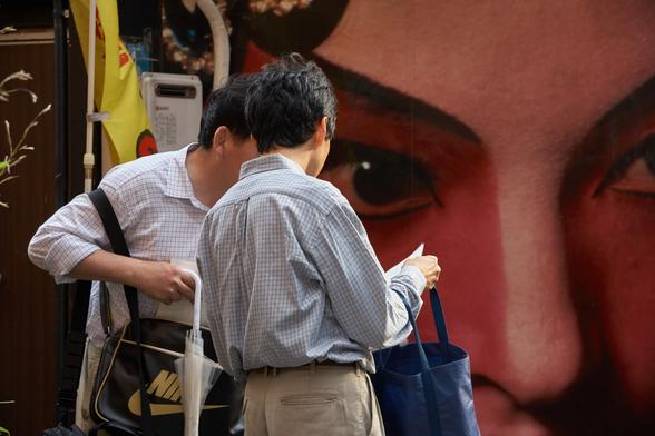 Two faceless salarymen share a moment looking at a printed page in front of a giant Kabuki billboard.