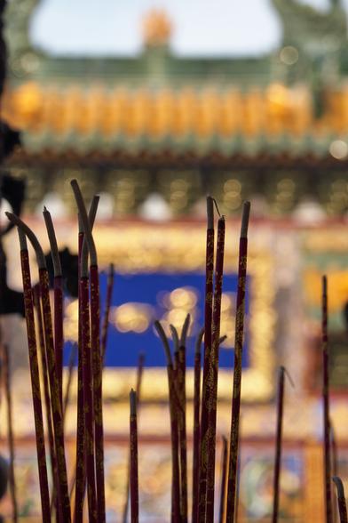 A closeup of burning incense sticks, offerings at the Yokohama Masobyo (Mazu Miao), the main colorful gate blurred behind them.