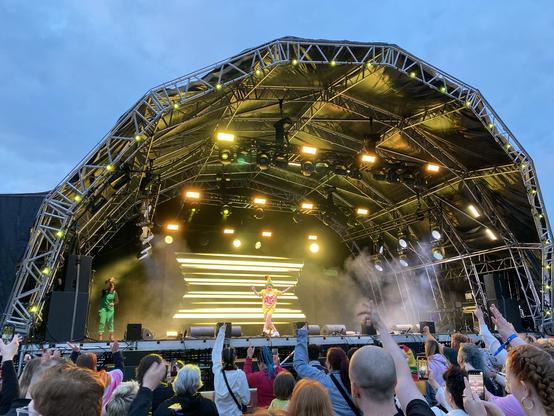 Outdoor concert with a GinnyLemon on stage and an audience cheering. The stage is lit with bright lights against a darkening sky.