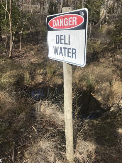 Warning sign next to a pond of water in the bush.
Sign should read DANGER DEEP WATER but has been altered to read DELI WATER.