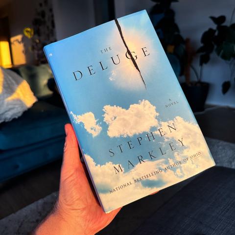 A hand holding a book titled "The Deluge" by Stephen Markley, with a cover featuring a blue sky and clouds.