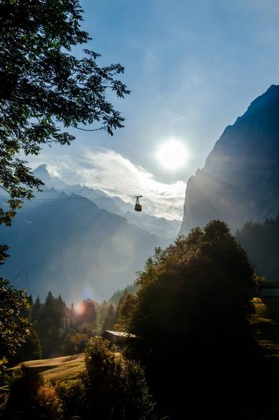 This image, captured by Hamburg wedding photographer Sturmsucht, depicts a serene mountain landscape bathed in soft morning or evening light. In the foreground, tall trees and lush greenery frame the scene, their leaves subtly illuminated by the sun. A single cable car is suspended in the air, moving across the expanse between two towering mountain peaks. The sun, positioned just above the horizon, casts a gentle glow, creating a hazy effect that envelops the distant mountains in a dreamy, ethereal light. The sky is clear, with only a few wisps of clouds adding texture to the blue expanse. The overall mood of the photograph is tranquil and contemplative, evoking a sense of peace and connection with nature.