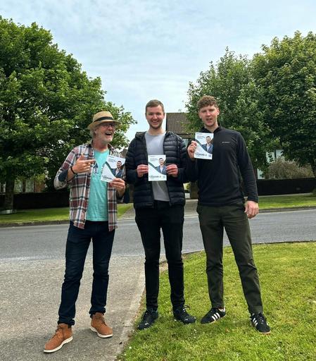 A photo of me holding my leaflet, a friend Fionn holding my leaflet, and his dad holding my leaflet, with trees and grass in the background in a housing estate.