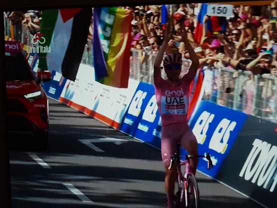 Tadej Pogacar crossing the finish line, 20th stage Giro D Italia 2024. Behind him are cheering fans behind the barriers and a rainbow flag with the letters PEACE on it, and the Palestinian flag above it.