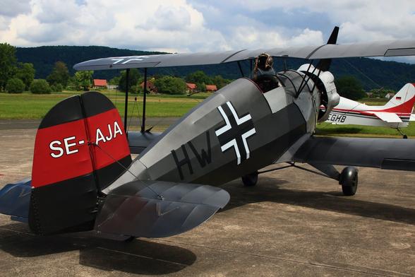 A single-seat open-cockpit "Bücker Jungmeister" vintage biplane with dark grey livery and Swedish registration, photographed from the right rear. Parts of a more modern white aircraft with red and black stripes in the background