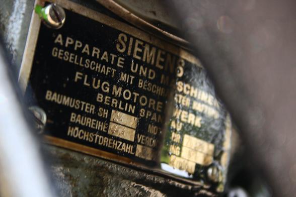 A type placque of a vintage radial engine reading "Siemens Apparate und Maschinen Gesellschaft mit beschränkter Haftung, Flugmotorenwerk Berlin Spandau", and some additional technical specifications, partially obscured by machine parts.