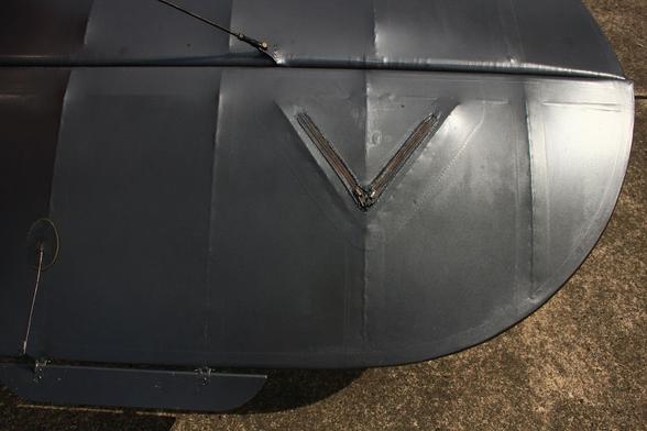 A close-up of the horizontal stabilizer and elevator of a grey-painted vintage aircraft. a triangular access flap is closed with what looks like regular zippers