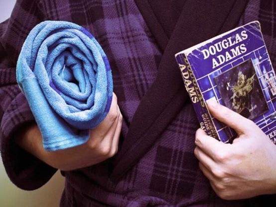 Persona en bata de cuadros con una toalla azul enrollada en su mano derecha y unlibro de Douglas Adams, también en azul, pero más oscuro, en su mano izquierda.