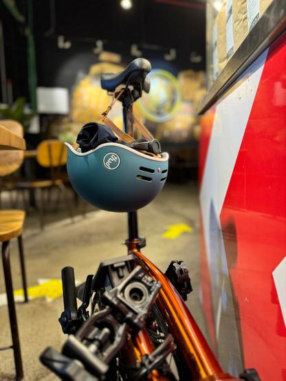 Brompton plegada con el sillín en alto y del que cuelga el casco con guantes y alguna cosa dentro.