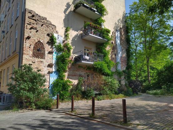 The side of an apartment building with a bit of very old brick wall incorporated into the newer wall and three balconies overgrown with vines seemingly randomly plonked onto the structure, giving the whole scene a rustic style - in the background more greenery and trees