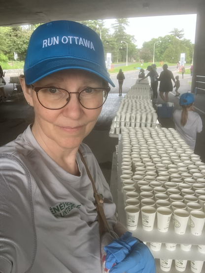 A woman wearing glasses, grey volunteer shirt and a blue ball cap stands besides tables full of compostable cups filled with water.