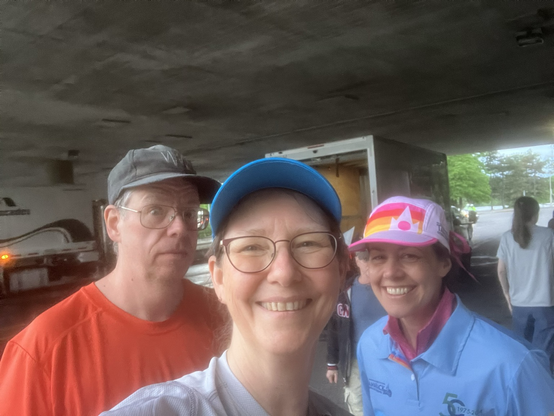 A man in a orange shirt looks concerned sarcastically, a woman in a blue cap and glasses smiles, and a woman in a white and pink cap and a volunteer leader shirt with a rainbow band smiles for a group selfie under an overpass