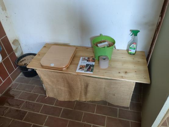 Eine Holzbank mit geschlossenem Toilettensitz, auf der Bank, neben dem Deckel ein Buch über Komposttoiletten, ein grüner Blecheimer mit Sägespänen zum bedecken der Scheisse und eine Toilettenpapierrolle. Um die Technik zu verstecken ist die Bank vorne mit einem Kartoffelsack zugehangen.