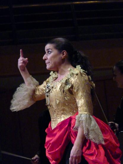 Cecilia Bartoli, seen in profile, wearing an elaborate bodice and ruched skirt, raises one finger and looks quizzically at the audience, pleading for 'one more'