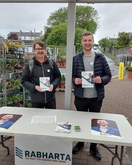 A photo of Killian and Eoghan standing behind a table set up with leaflets and posters for Killian's local election campaign.
