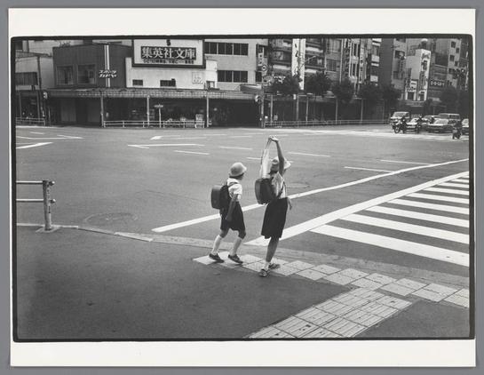 The image shows a city street scene with two people walking. It appears to be a black and white photograph, likely taken during daylight given the visibility of the surroundings. There is a car parked on the side of the street, a sign in the background that reads "Gekko," which might be a business or location name, and various buildings lining the street, indicating an urban setting. The individuals are casually dressed, one with a backpack. They are walking across what appears to be a pedestrian crossing with lines painted on the road for guidance. There is no text visible in this description provided.
