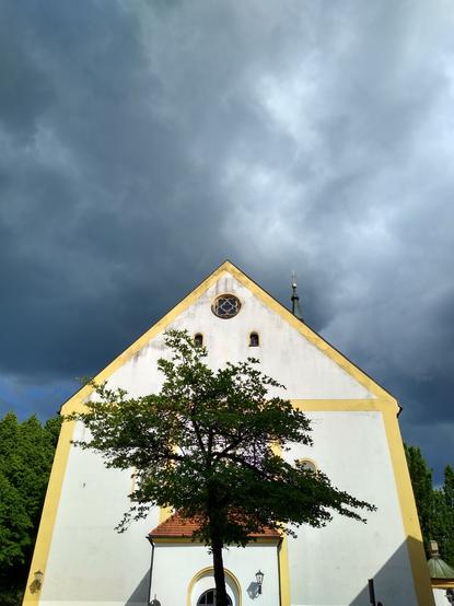 Kirche von vorne. Die Kirche ist weiß verputzt, teilweise gelb abgesetzt. In der Mitte vor der Kirche steht ein junger Baum. Die Sonne scheint auf die Kirche, der Himmel dahinter ist sehr dunkel bewölkt.
