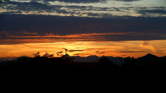 A shot towards west-north-west where the sun sets at the end of May at our geographic latitude. The sun has just set right behind our tallest mountain that is obstructed by an enormous construction crane. It was a beautiful sunset otherwise and the photograph shows stark contrasts between a pitch black silhouette of a suburban landscape, some tree canopies and distant hills and mountains, intensely orange horizon interspersed with broken clouds casting streaks of shadow, and the top half of the image are denser clouds reflecting darker orange light at its bottom and transiting towards cerulean blue of the twilight sky at their deck.