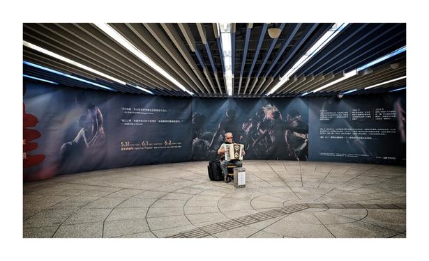 The image shows an elderly man playing an accordion in a subway station. He is seated on a small stool with his belongings next to him. The station's walls feature a large, dramatic mural promoting a theatrical performance at the National Theater. The mural depicts shadowy figures and includes text in Chinese and English about the event. The man is wearing sunglasses, and the ceiling above has modern, strip lighting. The overall atmosphere is quiet and reflective, with the musician being the focal point against the artistic backdrop.