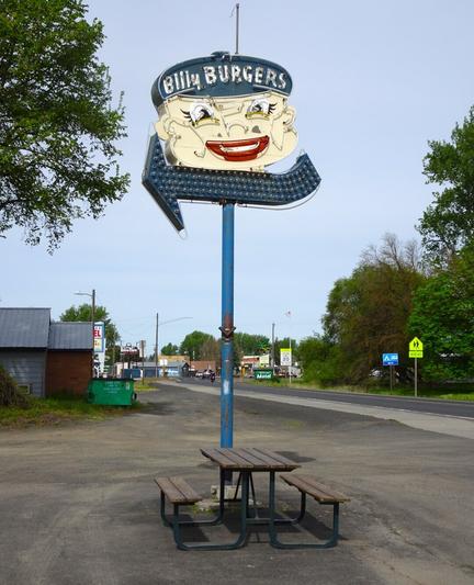 Billy Burgers sign in Wilbur, WA. The sign is neon with an arrow pointing leftward. The smiling boys face is a bit creepy with upturned nose and bright red lips. Beneath the sign is a single wooden picnic table at the roadside.