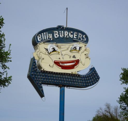 Close up photo of the Billy Burgers sign in Wilbur, WA. The sign is neon with an arrow pointing leftward. The smiling boys face is a bit creepy with upturned nose and bright red lips.