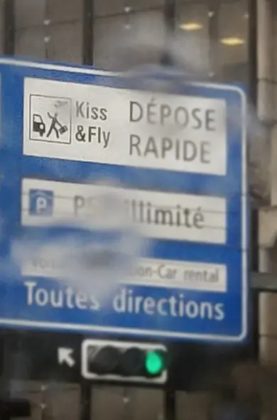 a street sign showing directions and a green traffic light. Everything is blurred by raindrops, except a part of the sign that spells "kiss and fly".