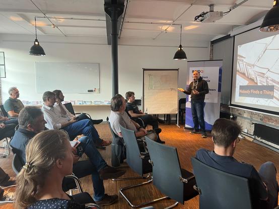 Das Bild zeigt eine Gruppe von Personen, die einer Präsentation zuhören. Der Redner Benjamin Wolf steht vor einer Leinwand, auf der "Nobody Finds a Thing" zu sehen ist. Die Zuhörer sitzen auf Stühlen in einem Raum mit industriellem Charme und weißen Wänden. Eine große weiße Tafel und ein Flipchart sind im Hintergrund sichtbar.