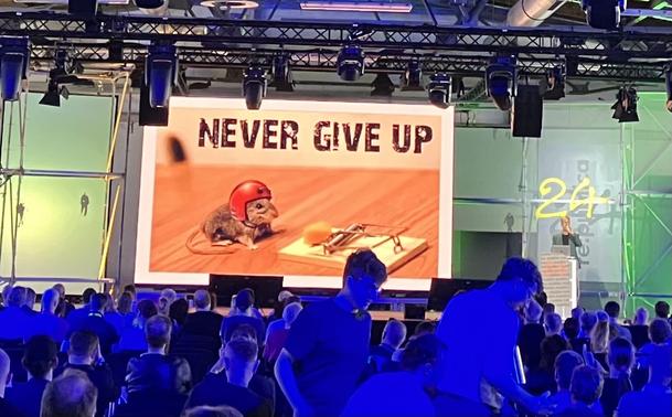 The screen on Stage1 showing a slide with a foto of a mouse and a mousetrap. The mouse is sitting in front of the trap wearing a red protective helmet. The headline of the slide says: Never give up.