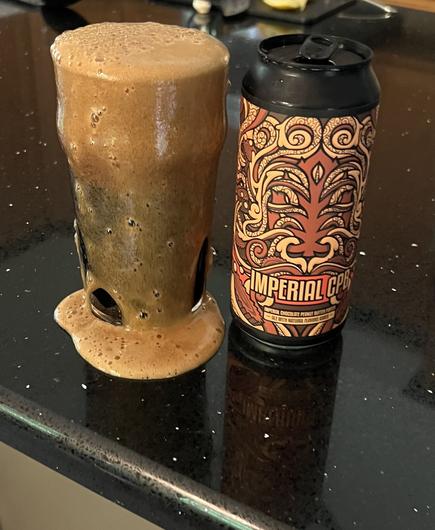 An overflowing pint glass of beer is coated in a deep tan head. The pint can of Lupulin Imperial CPB is to the left, its label in shades of brown, red, tan and orange. Peanut butter cups appear at the base.