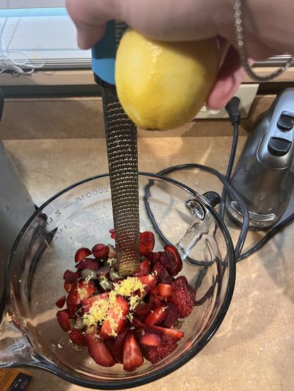 Photo of a hand holding a lemon and a microplane above the glass mixing bowl full of strawberries and rhubarb. Bits of lemon zest are sprinkled atop the fruits. A hand mixer is in the background.