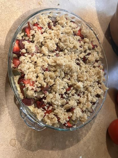 Photo of a glass pie tin full of dough, bits of fruit, and a crumble topping.