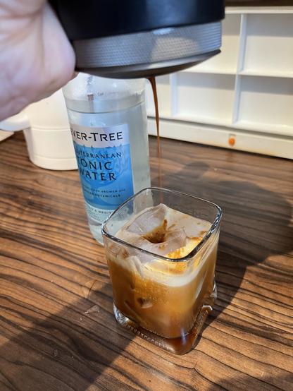 Ein Schuss Espresso, der über Eis in ein Glas gegossen wird, mit einer Flasche mediterranem Tonic Water im Hintergrund auf einer Holzoberfläche.