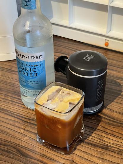 Ein Schuss Espresso, der über Eis in ein Glas gegossen wird, mit einer Flasche mediterranem Tonic Water im Hintergrund auf einer Holzoberfläche.